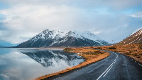 Zimná panoramatická fotografia cesty vedúca pozdĺž pobrežia jazera k vulkanickým horám. Vysoké skalnaté vrcholy pokryté vrstvou snehu zrkadliace sa na vodnej hladine. Pohľad vodiča na Islandský cestný okruh.